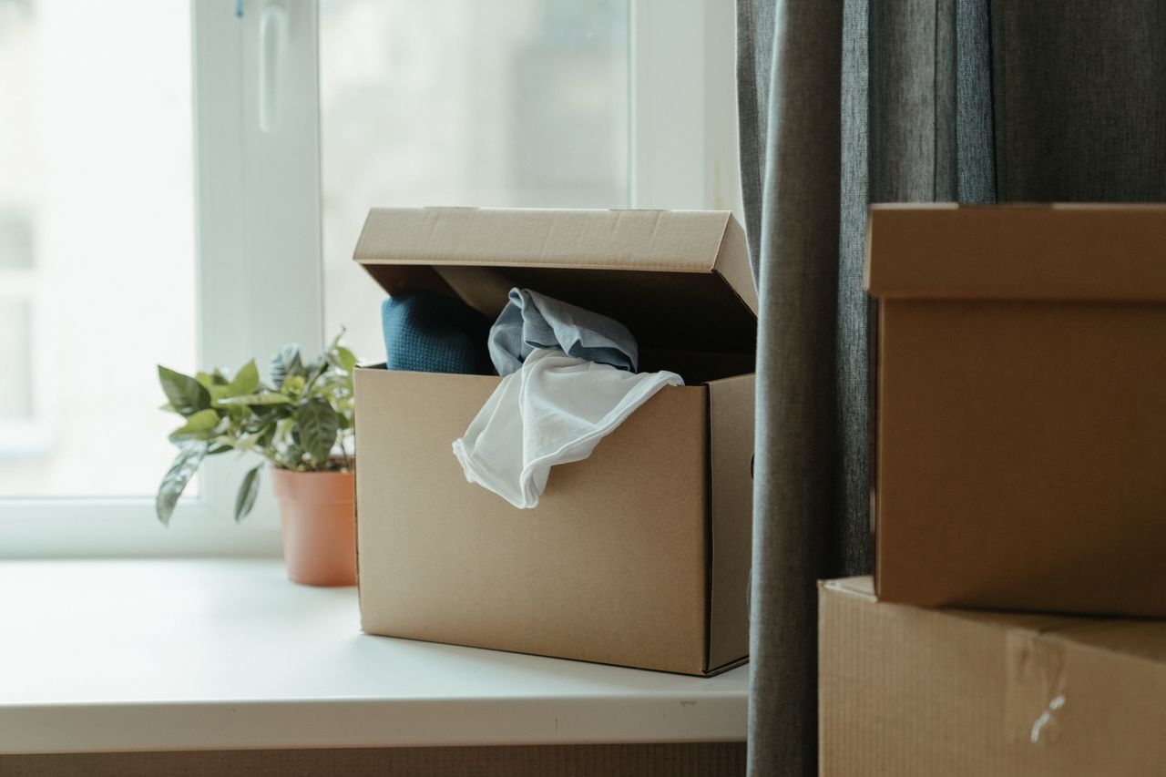 services-01 Open cardboard box with clothes by window, symbolizing moving or relocation.